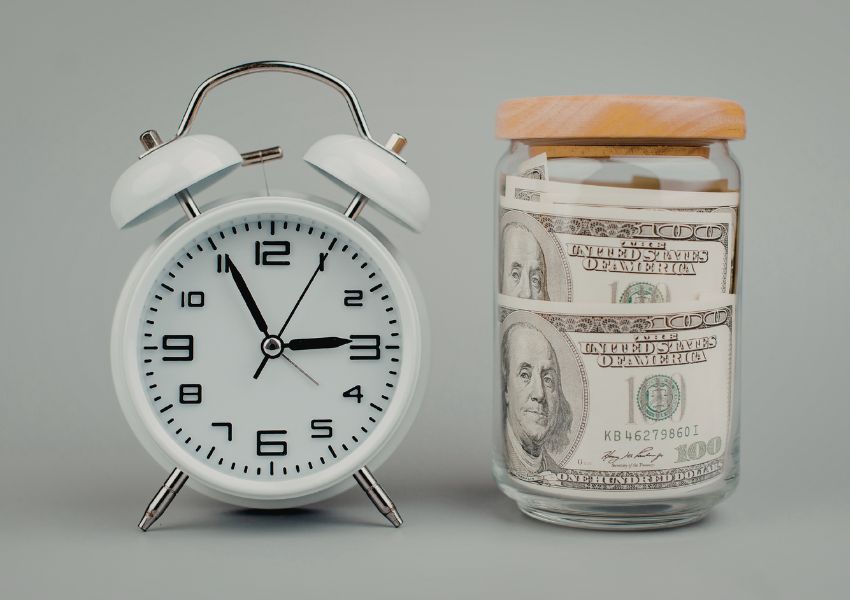A white alarm clock sits next to a glass jar filled with US dollar bills, symbolising the concept that time equals money.
