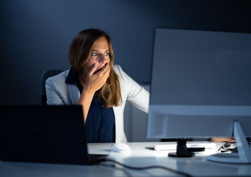 Woman sitting at a desk at night, covering her mouth in shock while staring at a desktop monitor, with a laptop and office equipment visible in a dimly lit workspace.