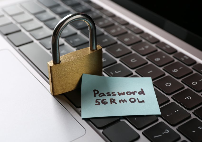 Close-up of a laptop keyboard with a padlock placed on top and a sticky note showing a handwritten password, highlighting weak password security practices.