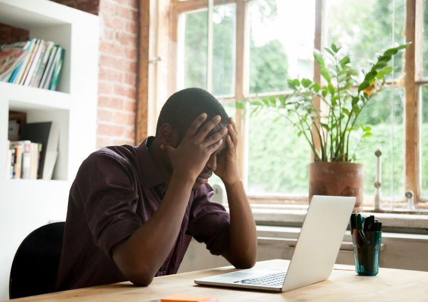 Man sitting at a desk with hands on his head while looking at a laptop in a bright office. Scene represents workplace stress, slow systems, or connectivity issues affecting productivity.