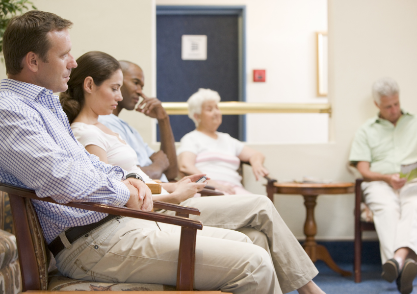 Patients seated in a clean doctor’s waiting room reading, using phones, and waiting for appointments.